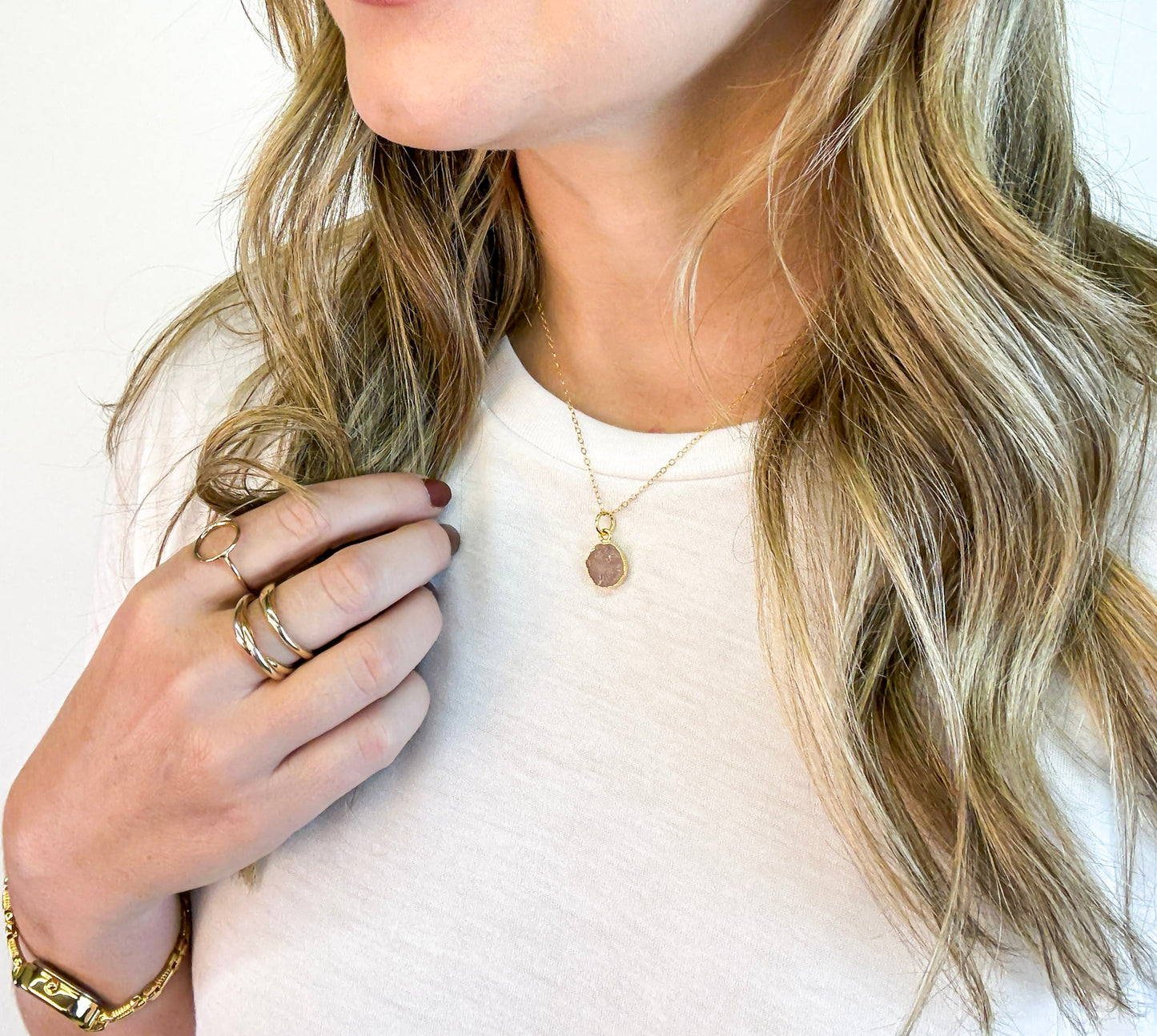 Close-up of a person wearing The Aurora Necklace by Studio Hill Designs. A handmade gold filled necklace with a pink stone pendant, white shirt, and gold jewelry.