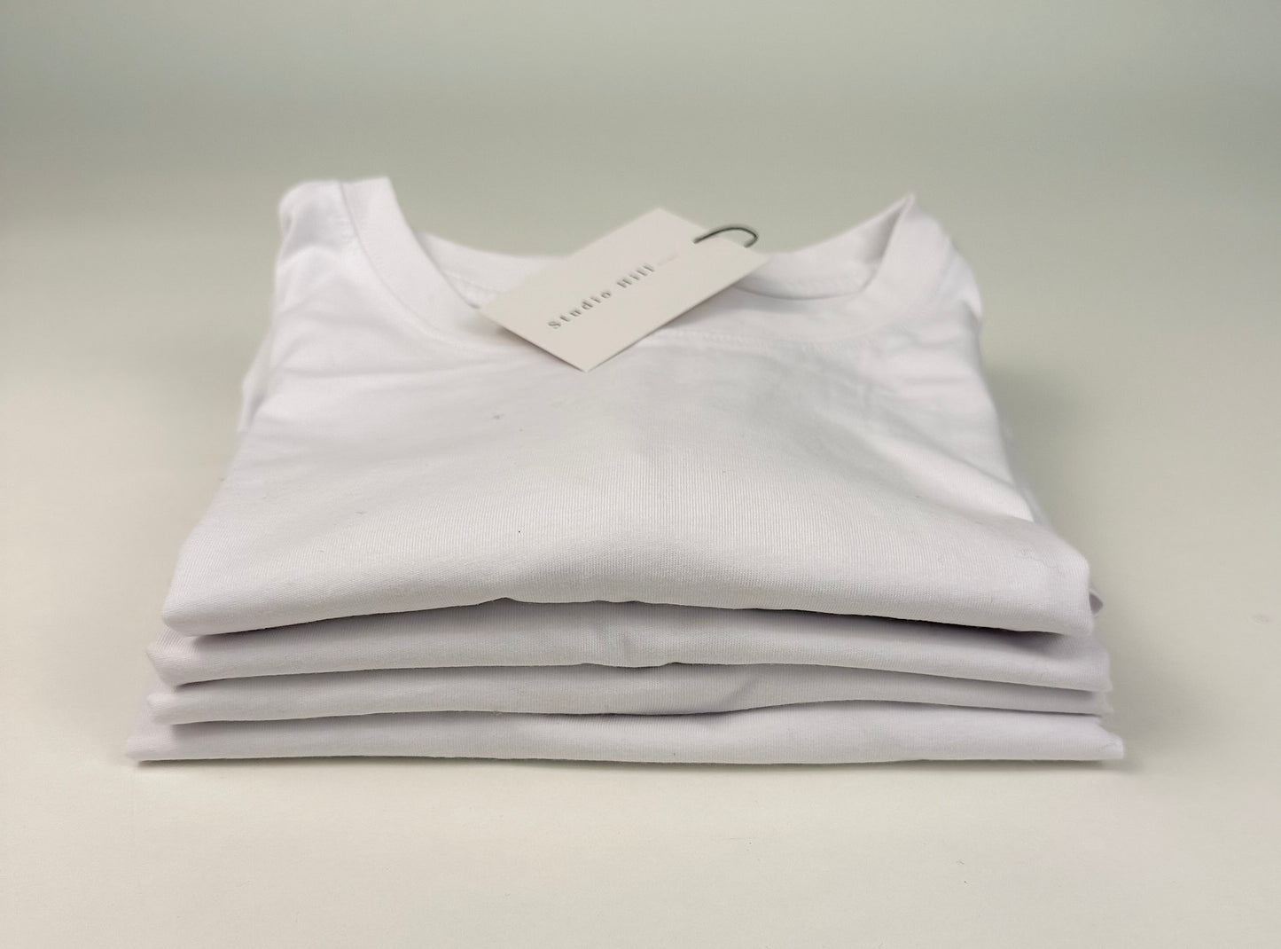 Several of The Essential Cotton Tee folded and stacked on white background by Studio Hill Designs.