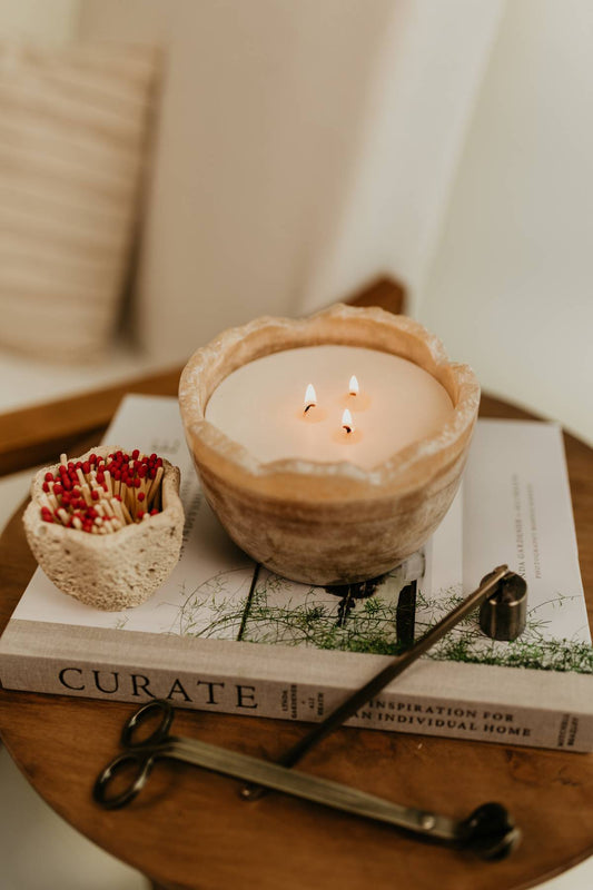 The Travertine Candle Bowl, a reusable stone bowl candle in earthy tones styled on a book with matches and a candle trimmer and snuffer on a wooden surface.
