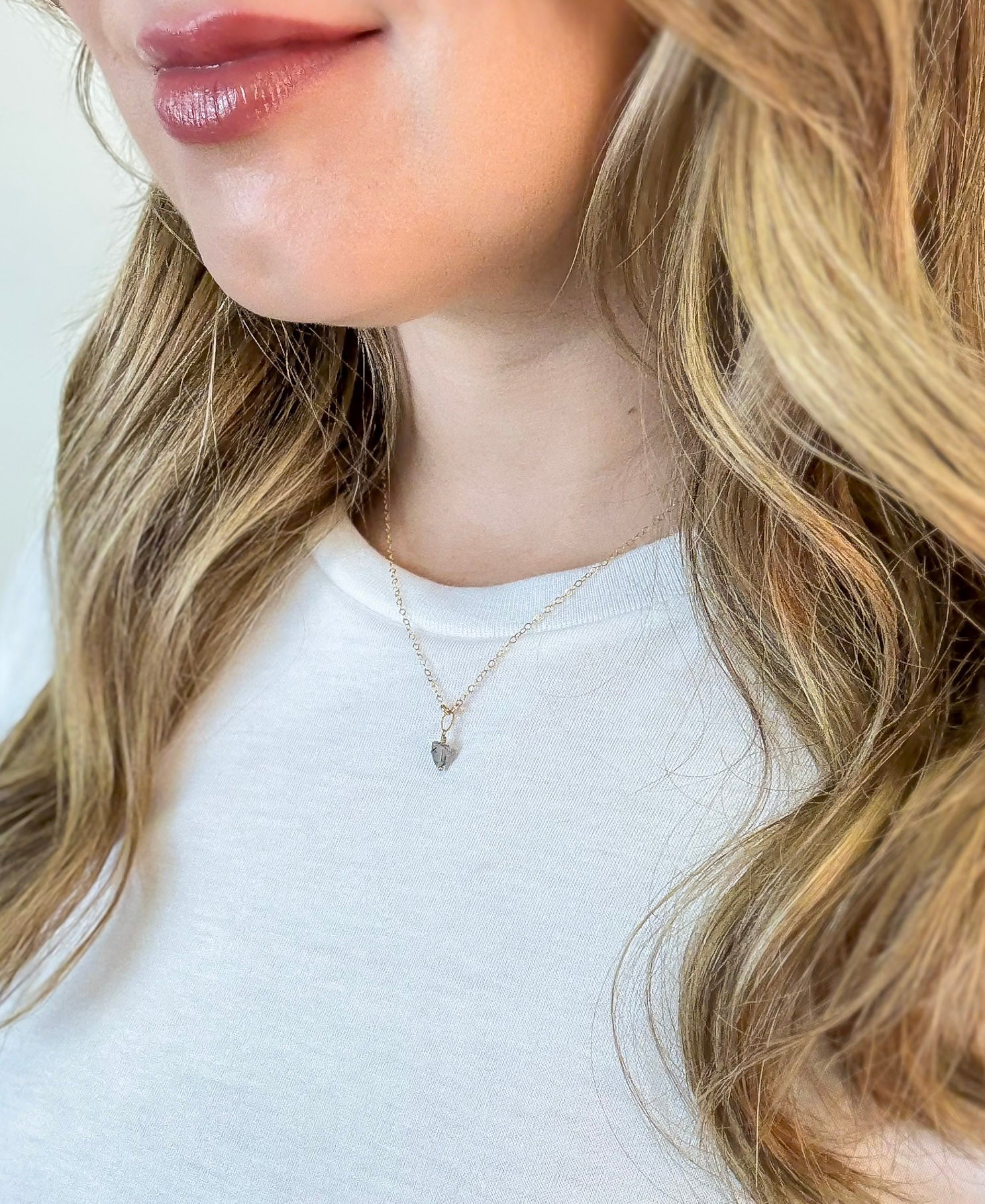 Close-up of a person wearing The Luna Necklace, a gold chain with black rutilated quartz pendant by Studio Hill Designs against a neutral background