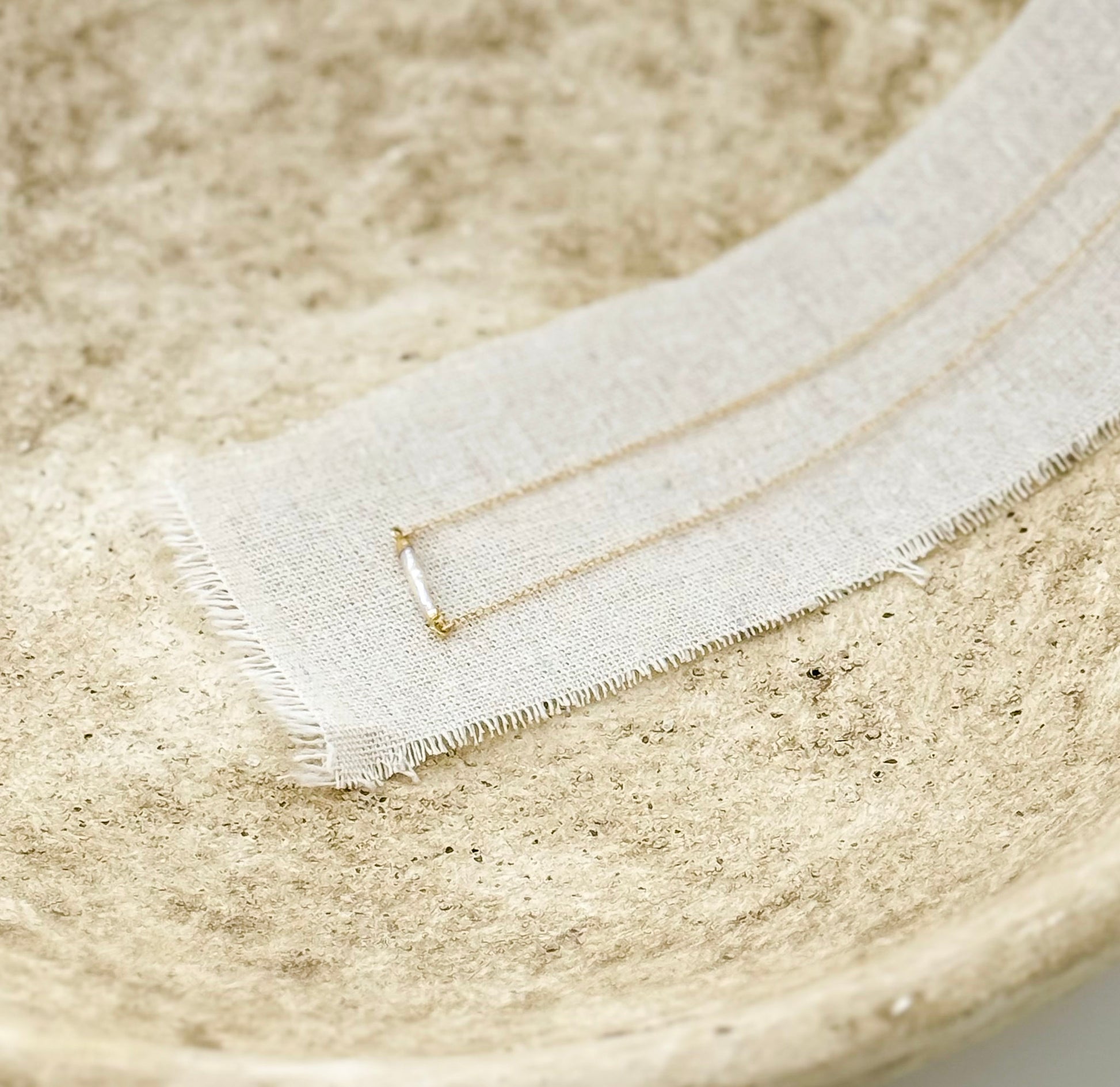 The Heirloom Necklace, a gold necklace with a pearl bar pendant, in a stone dish on a linen strip, by Studio Hill Designs.