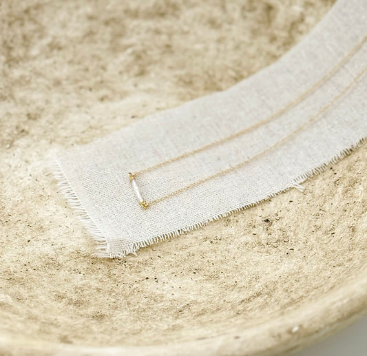 The Heirloom Necklace, a gold necklace with a pearl bar pendant, in a stone dish on a linen strip, by Studio Hill Designs.