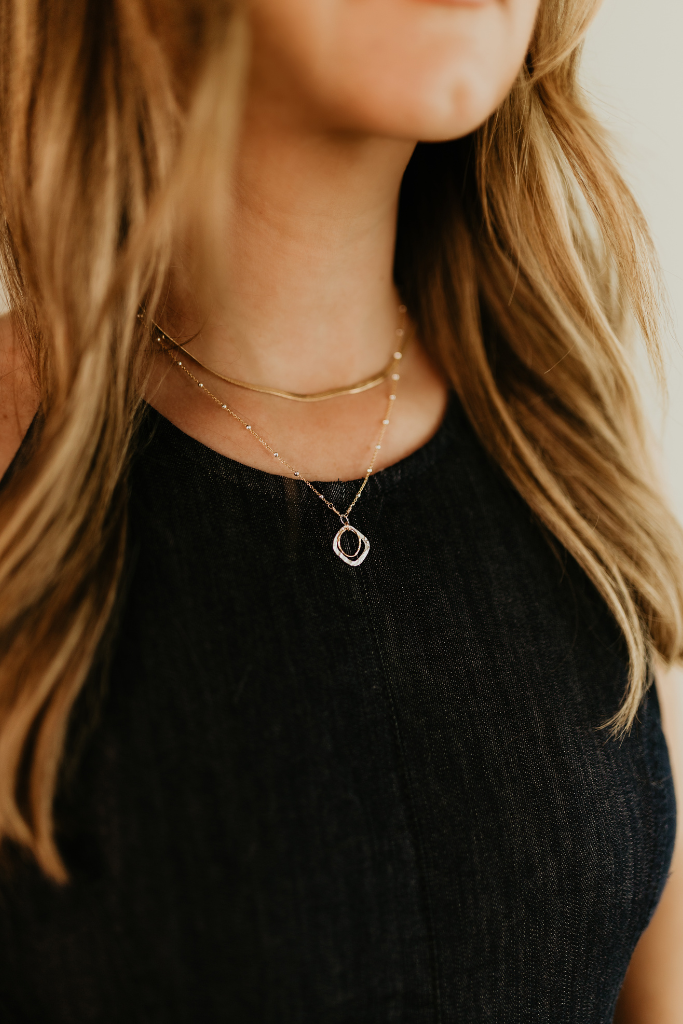Close up of person wearing The Serenity Necklace, a delicate gold and silver necklace by Studio Hill Designs with a small gold and silver hollow circle pendant against a neutral background