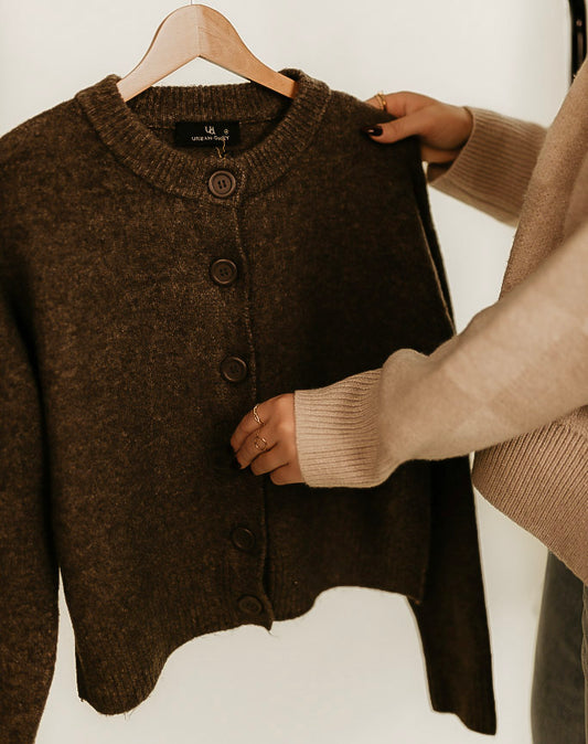 The Elise Wool Blend cardigan in brown on a hanger with a persons hands on the sweater showcasing the buttons.