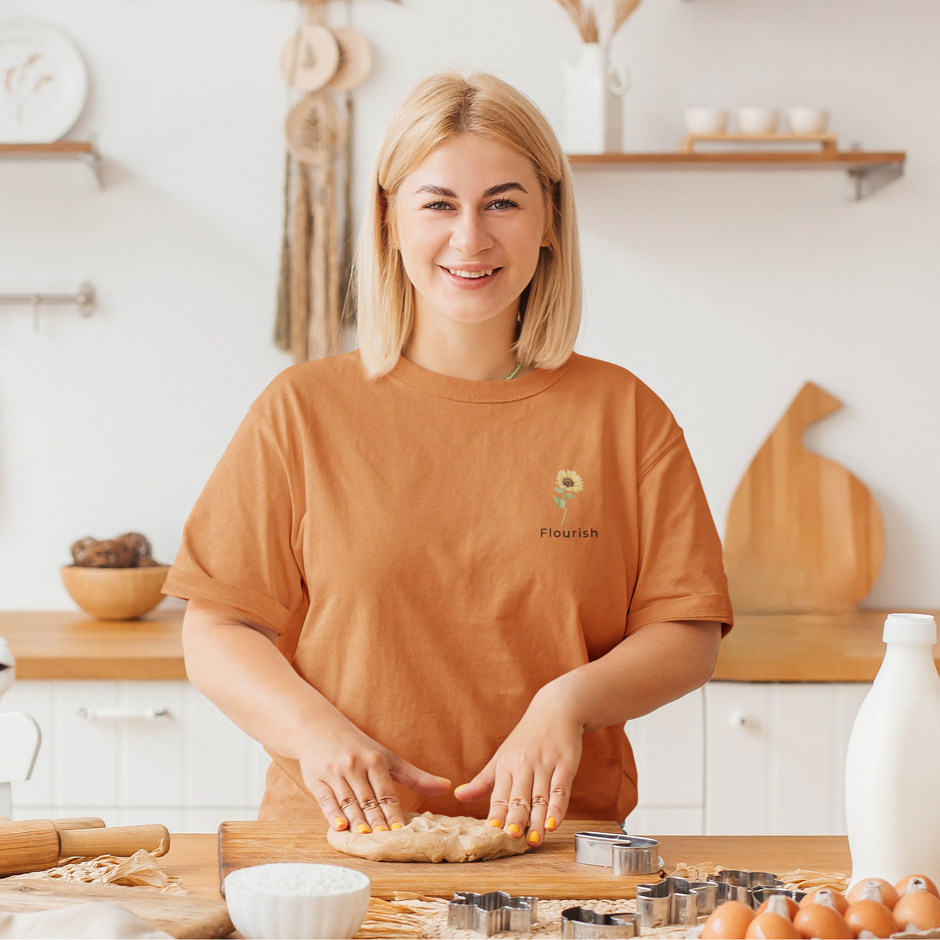 Woman in a kitchen wearing an orange t-shirt with small sunflower that says flourish underneath in the top corner by studio hill designs